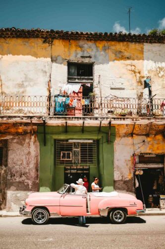 A pink vintage car parked in front of a weathered building in Cuba, with colorful laundry hanging on a balcony under the bright sun. (Photographer: Stephan Valentin)