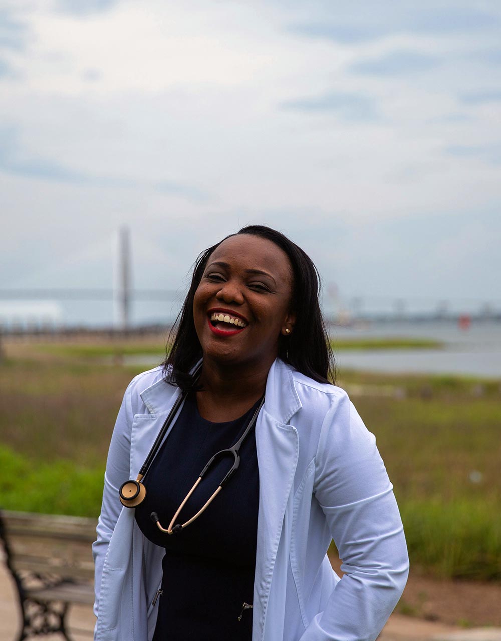 A young, Black female doctor with a stethoscope around her neck and white lab coat stands in front of a field.