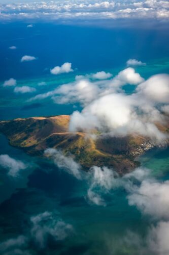 Aerial view of a tropical island in Fiji, partially covered by white clouds, surrounded by turquoise waters and a small coastal village. (Photographer: Janis Rozenfelds)