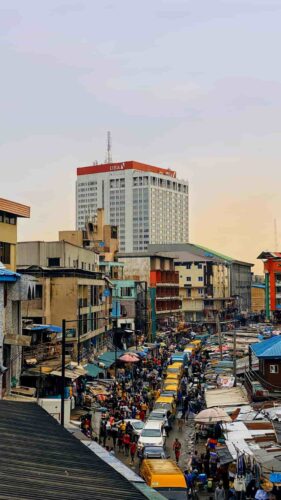 Busy street market in Lagos Marina, Nigeria, with yellow cars, vendors, and pedestrians, set against a backdrop of modern high-rise buildings. (Photographer: Namnso Ukpanah)