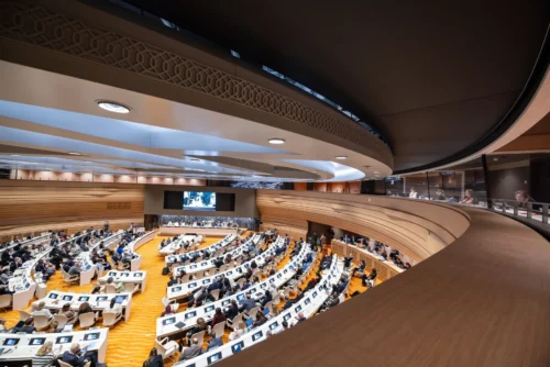 A wide-angle view of the World Health Assembly in session, showing delegates seated in a large, modern conference hall with curved rows and a central screen.