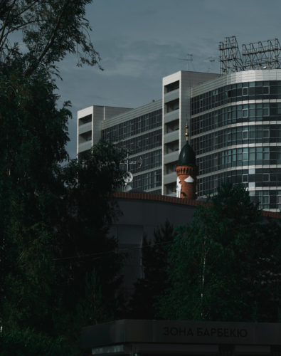 Modern multi-story building rises behind lush trees, with a distinctive minaret in the foreground, capturing a unique blend of contemporary design and traditional architecture.