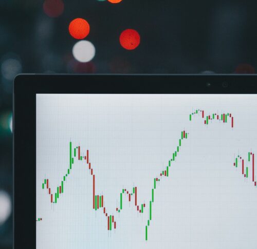 A computer screen displays a candlestick chart showing fluctuating stock or cryptocurrency prices, with blurred city lights in the background.
