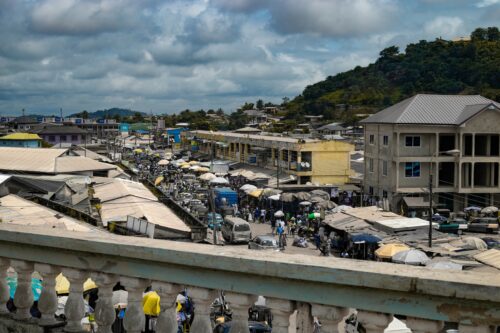 A busy urban market scene in a West African town, viewed from an elevated balcony.