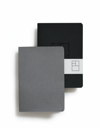 Two minimalist notebooks overlapping on a white surface. One is dark gray, the other black with a white band. The tone is clean and modern.
