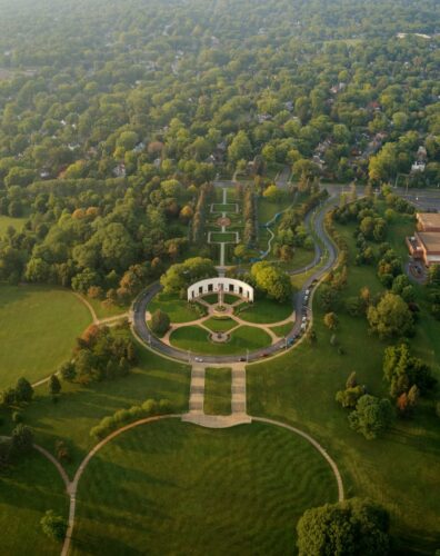 Aerial view of a lush park with a circular path leading to a central building, surrounded by sprawling lawns, gardens, trees, and nearby buildings.