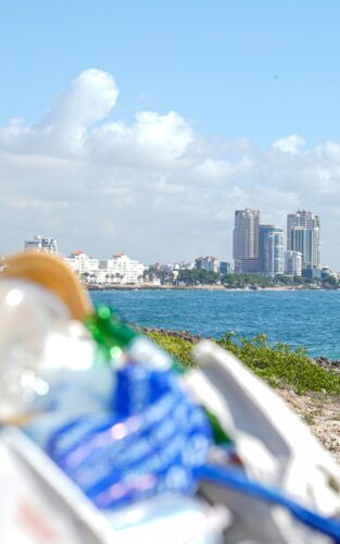 Plastic waste in the foreground with a coastal city skyline and ocean in the background, highlighting pollution near urban shorelines.