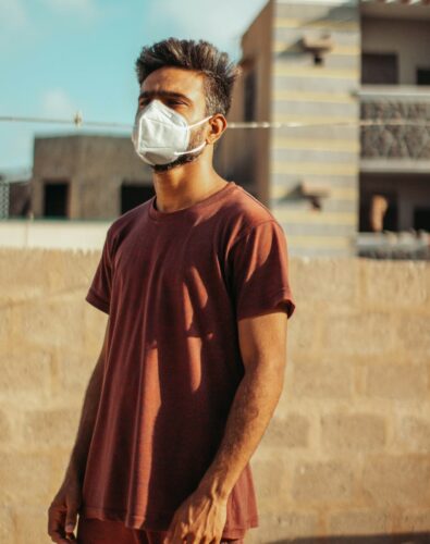 A man wearing a face mask stands outdoors in sunlight, with buildings and a stone wall in the background.