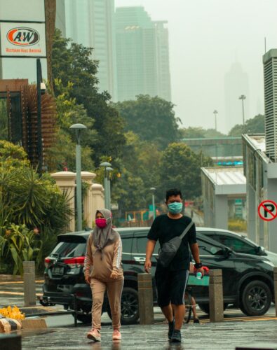 A man and woman wearing face masks walk on a rainy city sidewalk. The background shows wet roads, a parked car, and green trees, conveying a calm, overcast day.