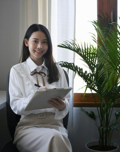 A woman in a white blouse sits indoors, holding a clipboard and pen, smiling at the camera with a potted plant and window in the background.