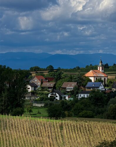 A tranquil village scene with a red-roofed church, quaint houses, and lush greenery, set against dramatic cloudy skies and distant mountains.