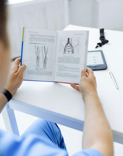 A person in blue scrubs reads a medical textbook at a white table. The book shows anatomical diagrams of legs. An e-reader and pen are nearby.