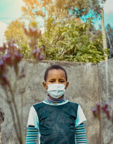 A young boy wearing a face mask stands outdoors against a concrete wall, surrounded by foliage. The lighting is warm.