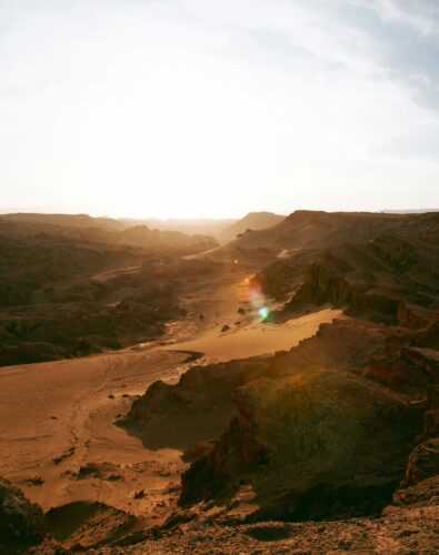 Sunset over a vast desert landscape, with rugged sand dunes and rocky formations. Warm sunlight casts a golden glow, creating a serene, tranquil atmosphere.