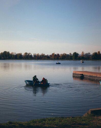 A serene lake scene with a small rowboat carrying two people in the foreground. Calm water, distant trees, and a wooden dock enhance the tranquil mood.