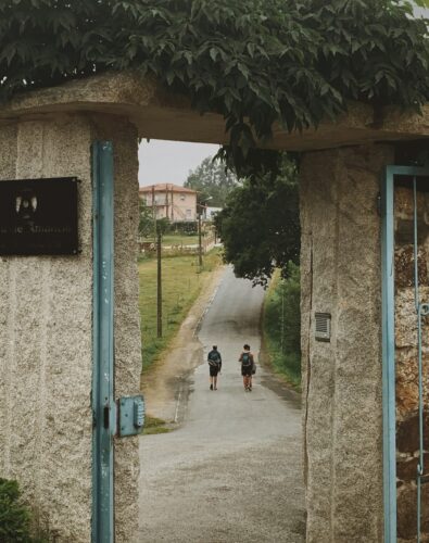 Two people with backpacks walk along a narrow, tree-lined road through an open stone gate. The mood is calm and rural, with a pink house in the distance.