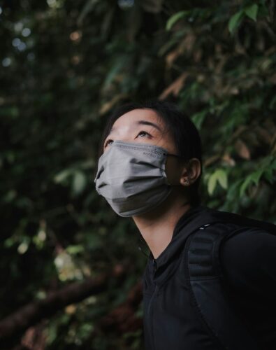 A person in a gray mask looks up amidst lush green foliage, wearing a black jacket. The mood is pensive and contemplative.