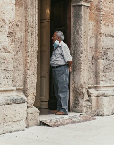 Older man stands in an ornate stone doorway, looking inside. A face mask hangs under his chin, his arms behind his back, conveying a contemplative mood.